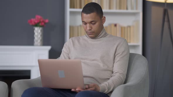 Middle Shot Portrait of Concentrated Young African American Man Sitting on Armchair Typing on Laptop