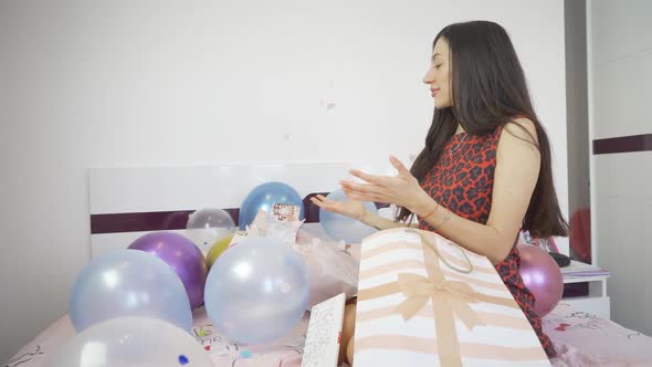 Side View of Woman Sitting on Bed with Colorful Balloons and Opens Confetti Bomb Birthday Card