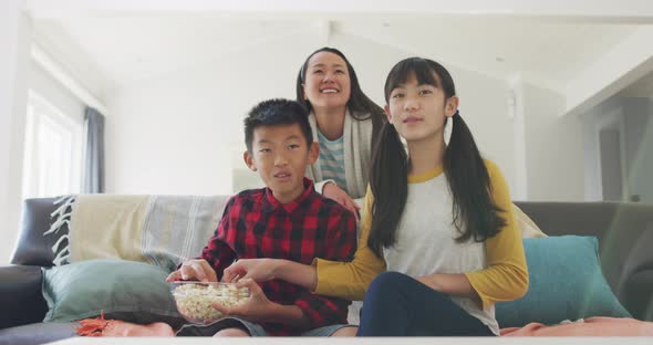Happy asian mother in living room watching tv with son and daughter sitting on couch eating popcorn