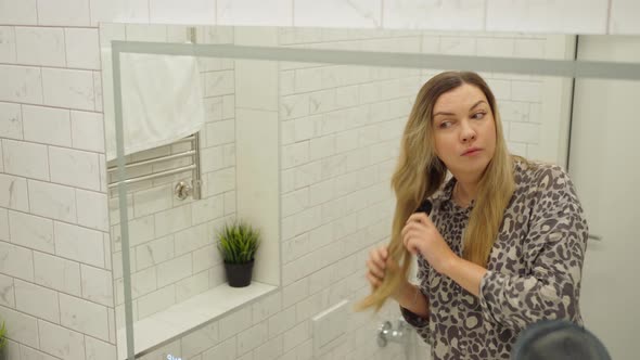 Young Woman Combing Her Long Blond Hair in Front of a Mirror in the Bath