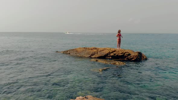 Girl Stands on Stone Midst Clear Blue Water Against Boat