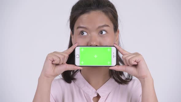 Face of Young Happy Asian Businesswoman Thinking While Showing Phone