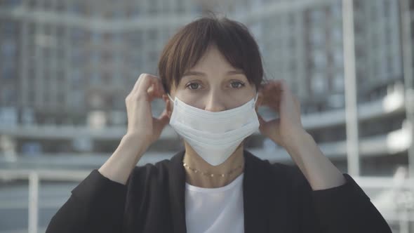 Close-up Portrait of Young Caucasian Woman Putting on Covid Face Mask and Looking at Camera. Serious