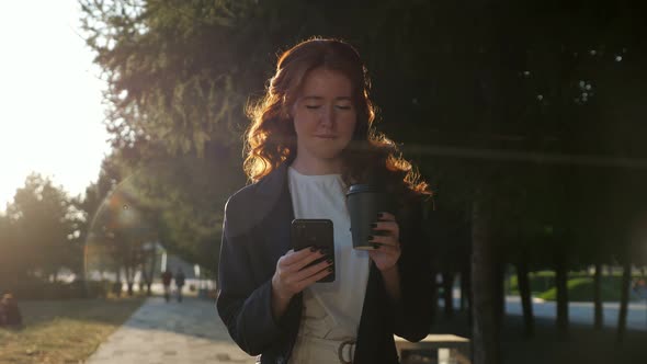 Positive Young Woman Holds Cup and Mobile Phone in City Park