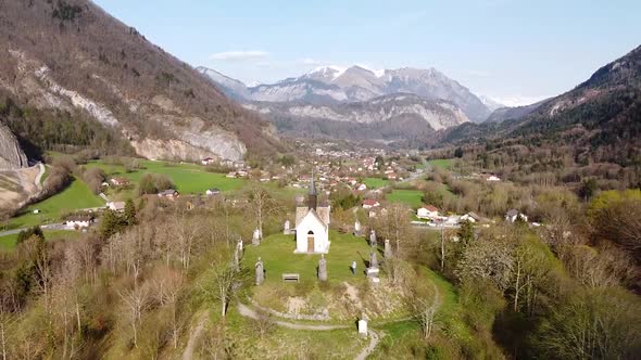 Drone Shot (facing forwards,ing up and back) of a Chapel on top of a Hill in front of Alpine Mountai