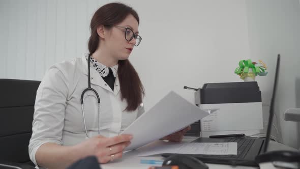 A Young Woman Doctor in Glasses Meets with a Patient Virtually Using Video Call Technology Holds the