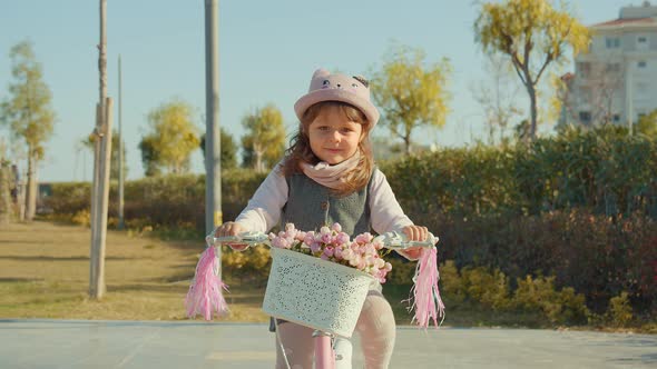 Little Cute Girl On A Pink Bicycle With Flowers Rides In The Park