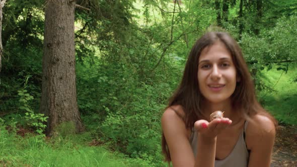 A woman on a hike in a green forest holding a snail and blowing it a kiss to the camera.