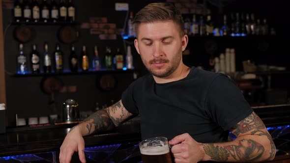 A Bearded Young Man with Tattoos Drinking Beer at Bar or Pub