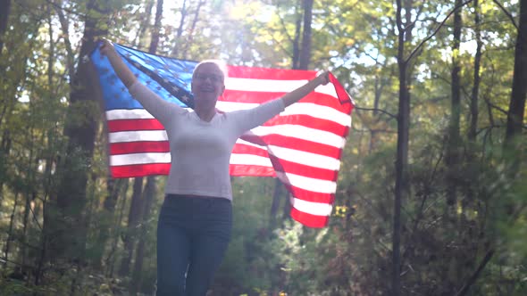 Backlit, pretty blonde woman walking through a forest holding a flag up in the air behind her.