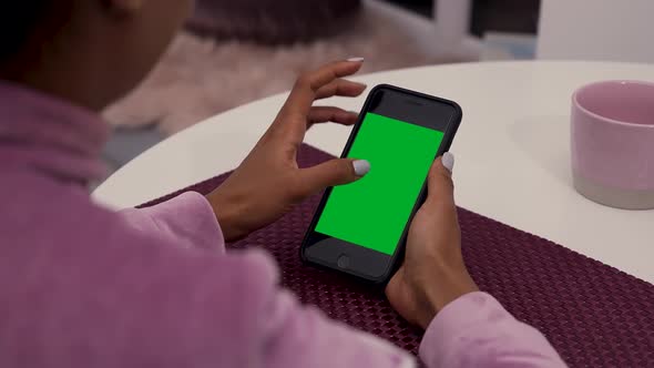 Young African American Woman Sits at Table in Room and Uses Smartphone with Green Screen Chroma Key