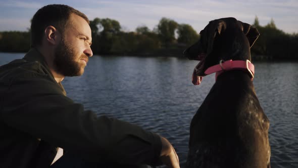 Young Guy Sitting with Dog on the Pier and Admiring the Beauty of the Lake