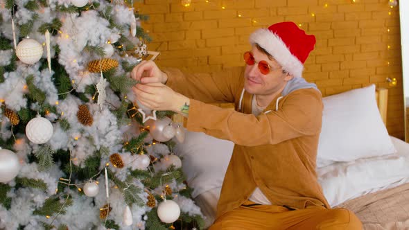 Man in Santa Hat and Fashionable Glasses Sitting on Bed and Decorating Christmas Tree