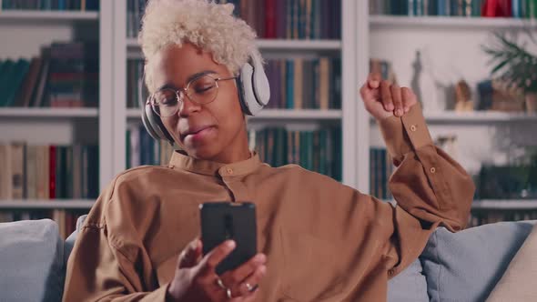 African Woman Relaxing on Sofa Holds Smartphone Listens Song Through Headphones