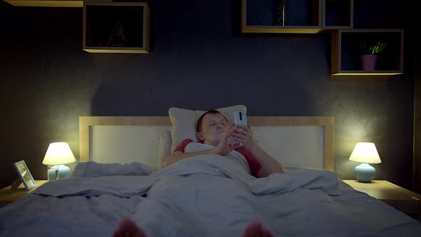 Man Lying in Bed at Night Using His Cell Phone Browsing the App Bedroom Interior Front View