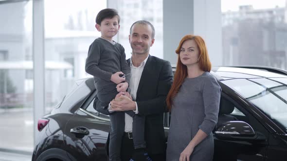 Happy Caucasian Family Standing in Car Dealership, Looking at Camera and Smiling