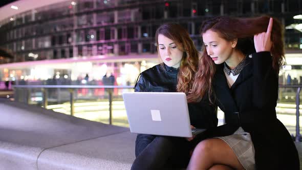 Two young beautiful caucasian women outdoor in the night using computer, discussing