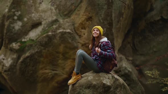 Happy Inspired Woman Hiker in Checkered Jacket and Beanie with Backpack Sits on Edge of Cliff and