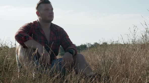 Lonely Young Man In Nature Sits In Tall Grass And Looks Around. Sad Man In The Field.