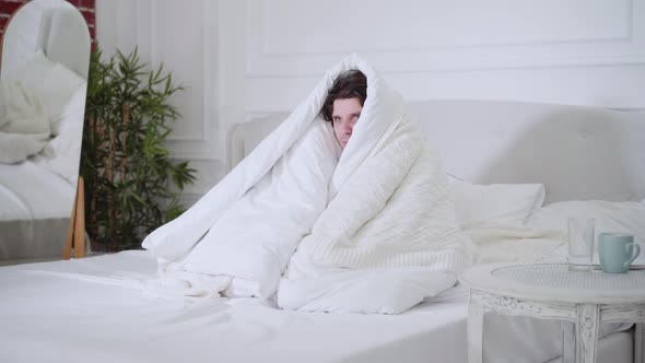 Ill Young Man Having Fever Sitting on Bed Wrapping in White Duvet
