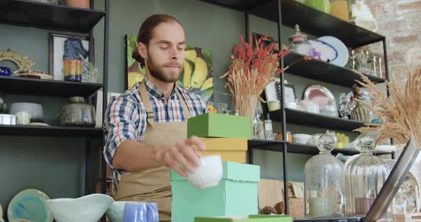 Bearded Man in Casual Clothes and Apron Sitting at Workplace in Beautiful Gift Shop