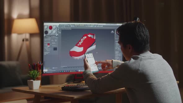 Asian Male Footwear Designer Looking At Photos On Smartphone While Designing Shoe On A Desktop