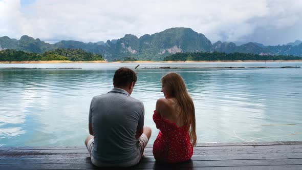 Man Teaching Woman to Beat Showing on Himself on Lake with Green Mountains