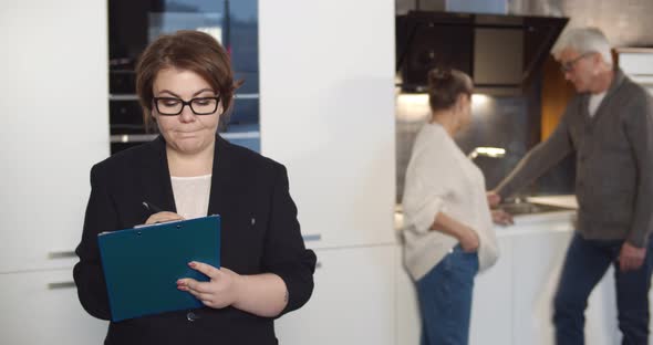 Woman Realtor Filling in Document on Clipboard While Aged Couple Examining Kitchen on Background