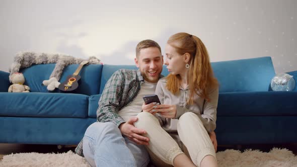 Couple Use Smartphone Device While Sitting on a Couch in the Cozy Apartment