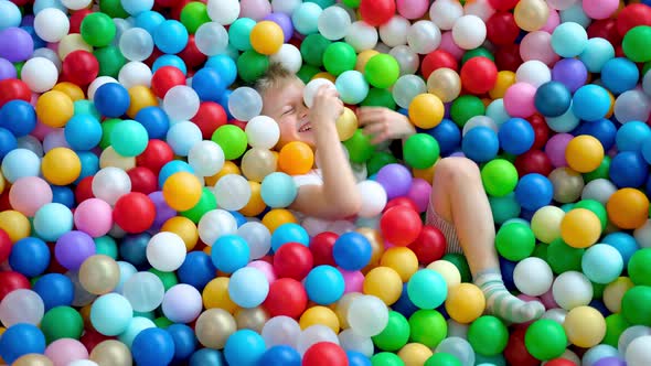 Blonde Little Boy Scatters Balls in Different Sides Lying on Multi Colored Plastic Balls in Big Dry