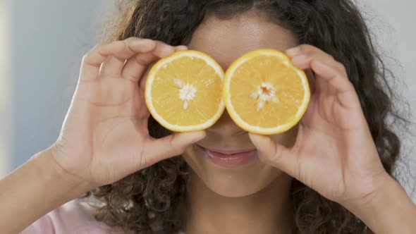 Portrait of cheerful biracial woman applying halves of orange to eyes, optimism