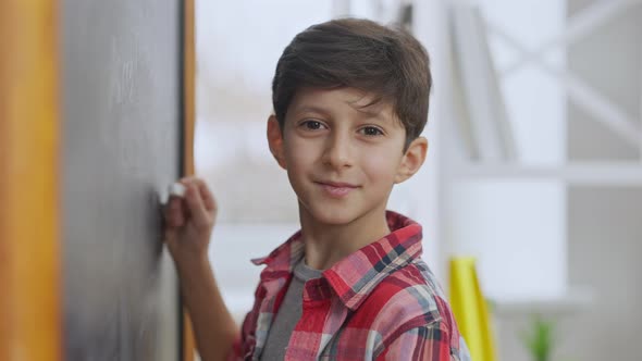 Closeup Portrait of Cute Middle Eastern Schoolboy Writing on Chalkboard Looking at Camera and