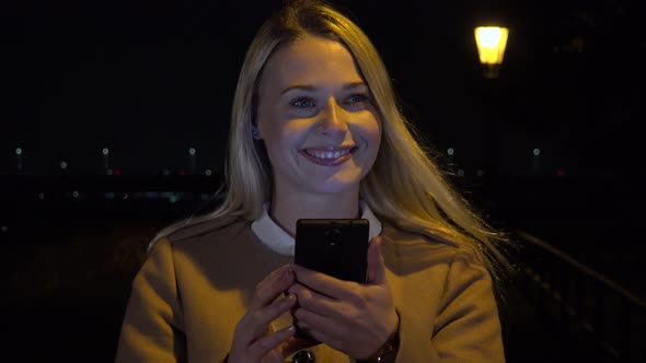 A Young Beautiful Woman Works on a Smartphone in an Urban Area at Night, Looks Up and Smiles