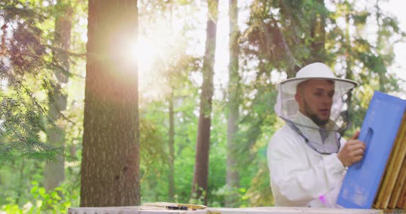 Young Male Bearded Beekeeper in White Protective Suit Takes the Section of Hive with Beehive Frames