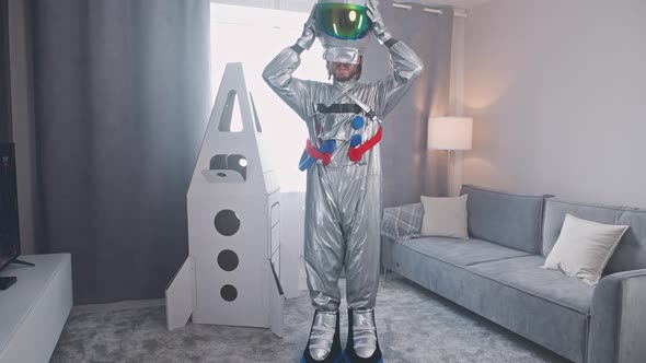 Middleaged Man in an Astronaut Suit Stands in Living Room Next to a Cardboard Model of a Space