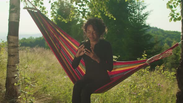 Charming African American Woman Traveler in Hammock Browsing Online on Phone in Mountain Top