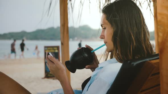 Side View Shot of Young Relaxed Freelancer Woman with Exotic Drink Watching Videos on Smartphone