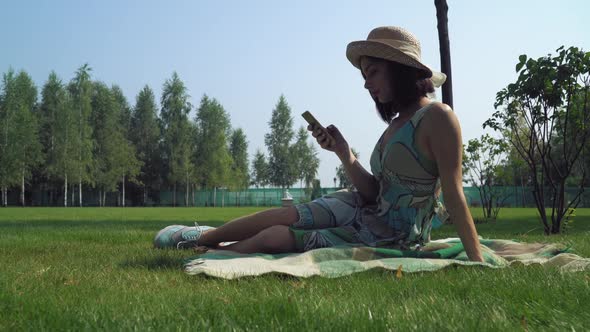 Young Brunette Girl Sitting on Blankets in the Park and Gaining a Message on the Phone