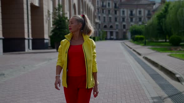 a Woman in Red Clothes and a Yellow Jacket Walks Along the Path Between the Park and the City