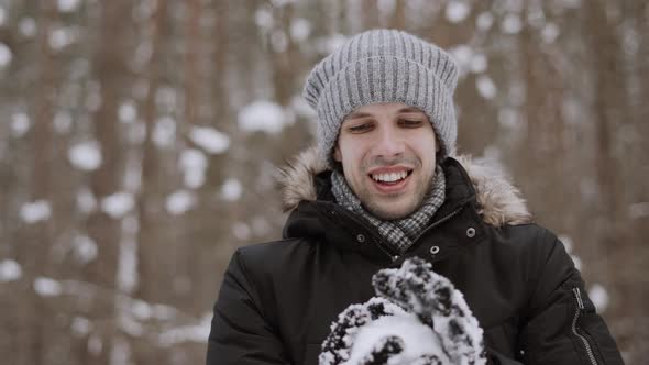 Portrait of Caucasian Young Man with Smile in Hat Sculpts Snowball in Gloves