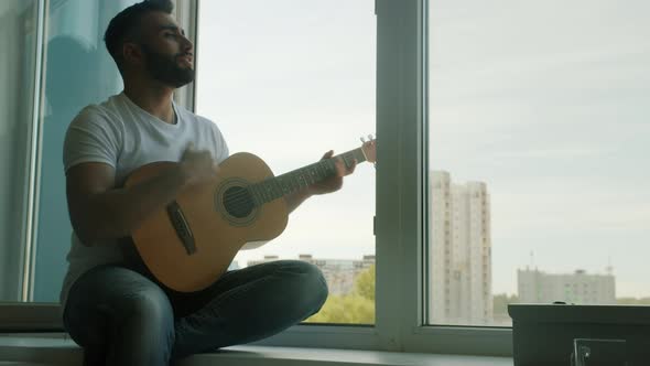Handsome Young Man Playing Guitar and Singing Having Fun Sitting on Window Sill at Home
