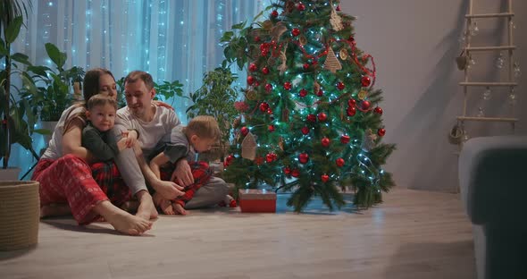 A Family with Two Children Sit Together at the Christmas Tree and Cuddle