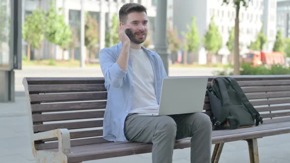 Young Man Talking on Phone and Using Laptop While Sitting on Bench