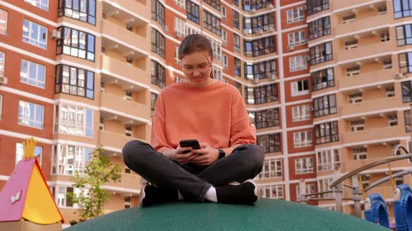 A Smiling Teenage Girl with Glasses is Sitting in the Courtyard with a Phone