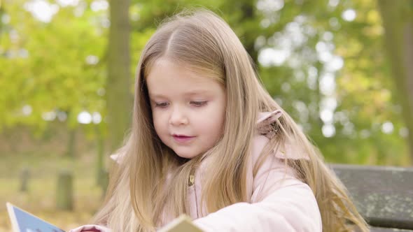 Cute Little Caucasian Girl Reads a Picture Book Out Loud with a Smile As She Sits on a Bench in Park