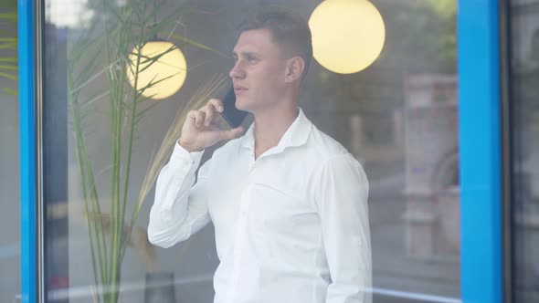 Portrait of Successful Caucasian Man Talking on the Phone Behind the Window. Young Confident Guy in