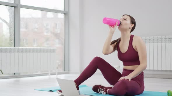 a Young Woman Drinks Water in a Burgundy Tracksuit with Laptop on a Mat in a Gym