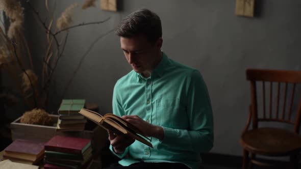 Medium Shot of Focused Young Man Flips Through Pages of Old Frayed Book Sitting in Chair in Dark
