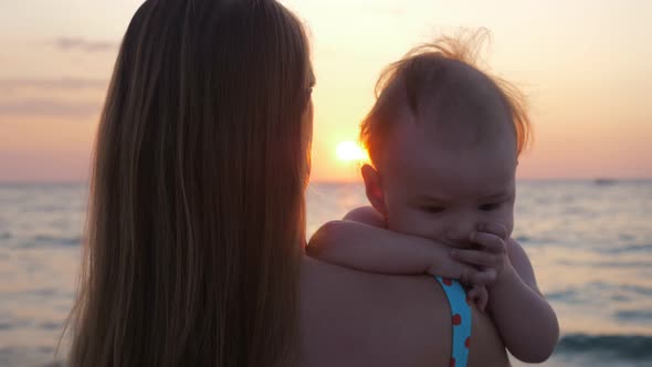 Mother Holds Baby in Her Arms While Looking at Sunset By the Sea or Ocean Shore.
