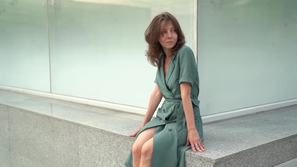 Sexy Middle-aged Woman Is Posing Outdoors, Sitting on Stone Bench at Modern City